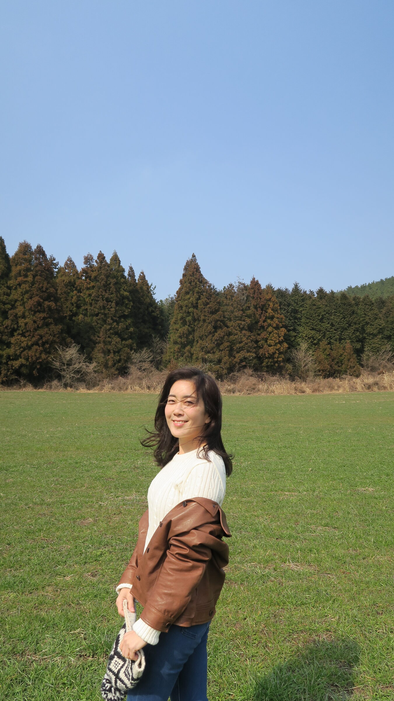 Jamie (Hee Jung) Lee standing in a grassy field in Jeju, wearing a white sweater and brown leather jacket